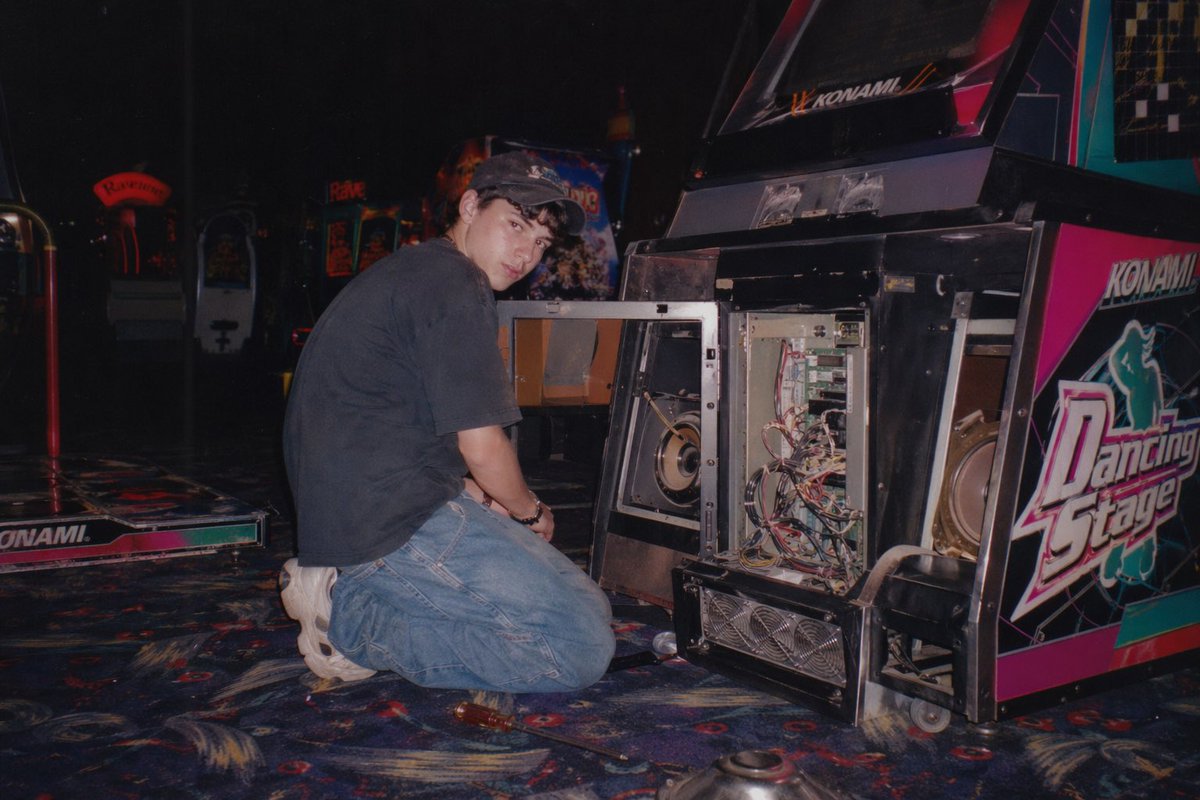 A vintage, late 90s amateur flash photograph of a young man repairing an arcade… — AI-generated image by @nicdunz using GPT Image 2