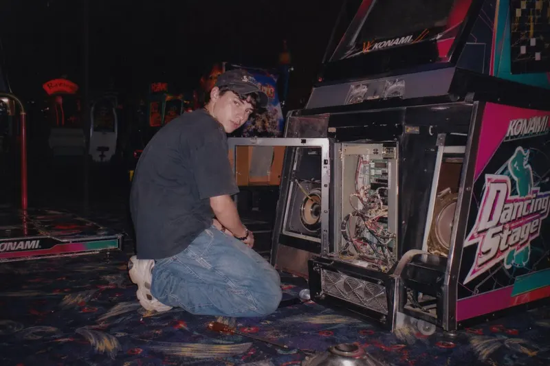 A vintage, late 90s amateur flash photograph of a young man repairing an arcade… — GPT Image 2 prompt by @nicdunz